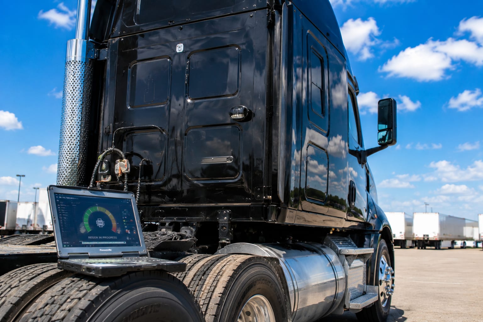 Rugged diagnostic laptop propped on a heavy truck showing a DPF regeneration in progress under a bright blue sky